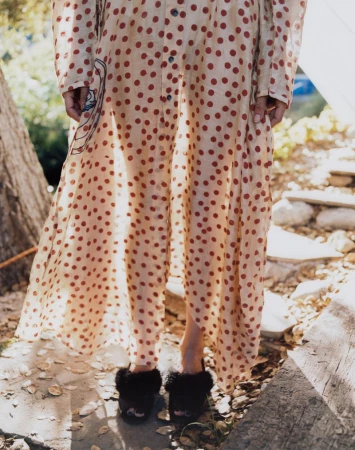 Model wearing a sheer polka dot dress and black furry slides standing outdoors on a stone path.