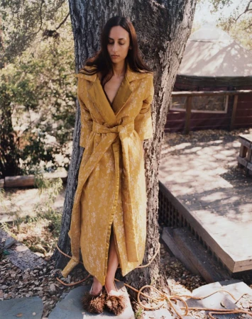 Model leaning against a tree trunk wearing a long yellow patterned coat and fluffy slip-on shoes outdoors.