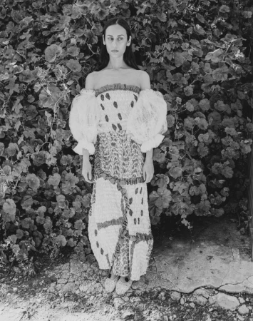 Model standing in a long patterned dress with voluminous puff sleeves against a dense background of dark foliage.