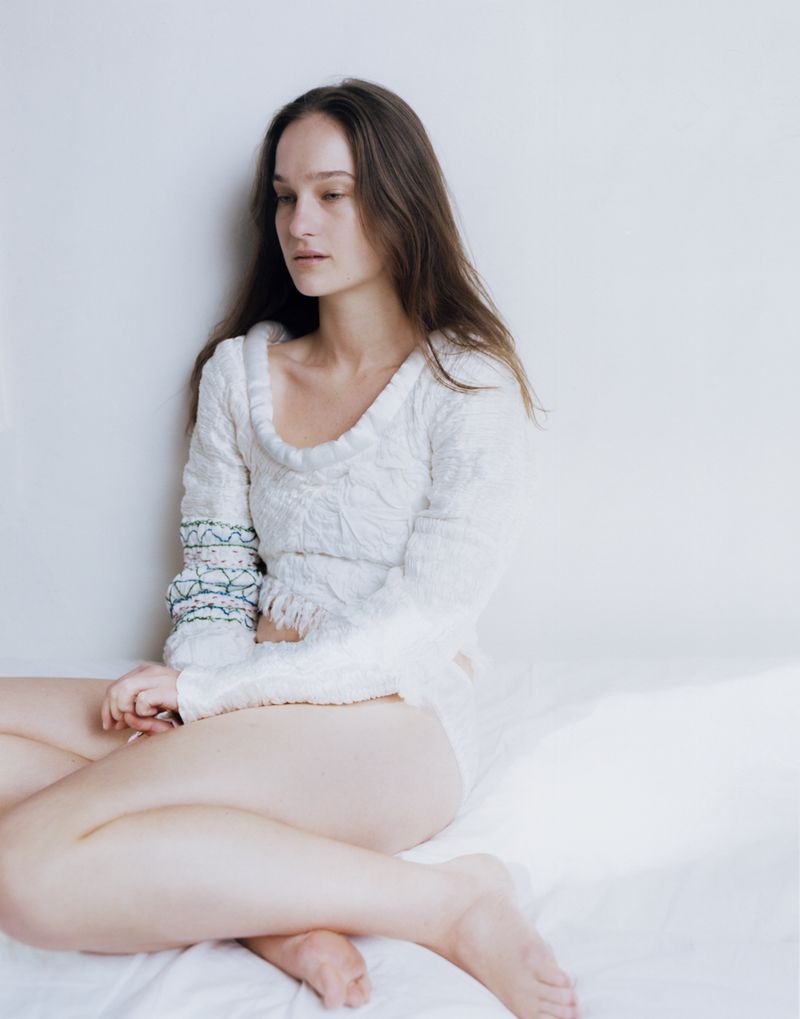 Model with long brown hair seated against a white background wearing a textured white sweater with embroidered sleeve detail