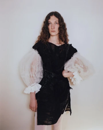 Model with long brown curly hair wearing a black dress with voluminous sheer white puffed sleeves against a plain background