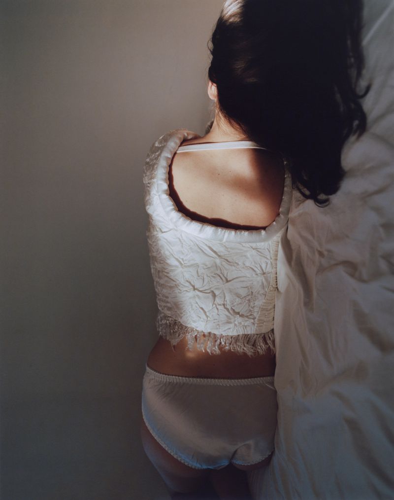 Overhead view of a woman with dark hair wearing a white textured top and white briefs seated on white bedsheets.