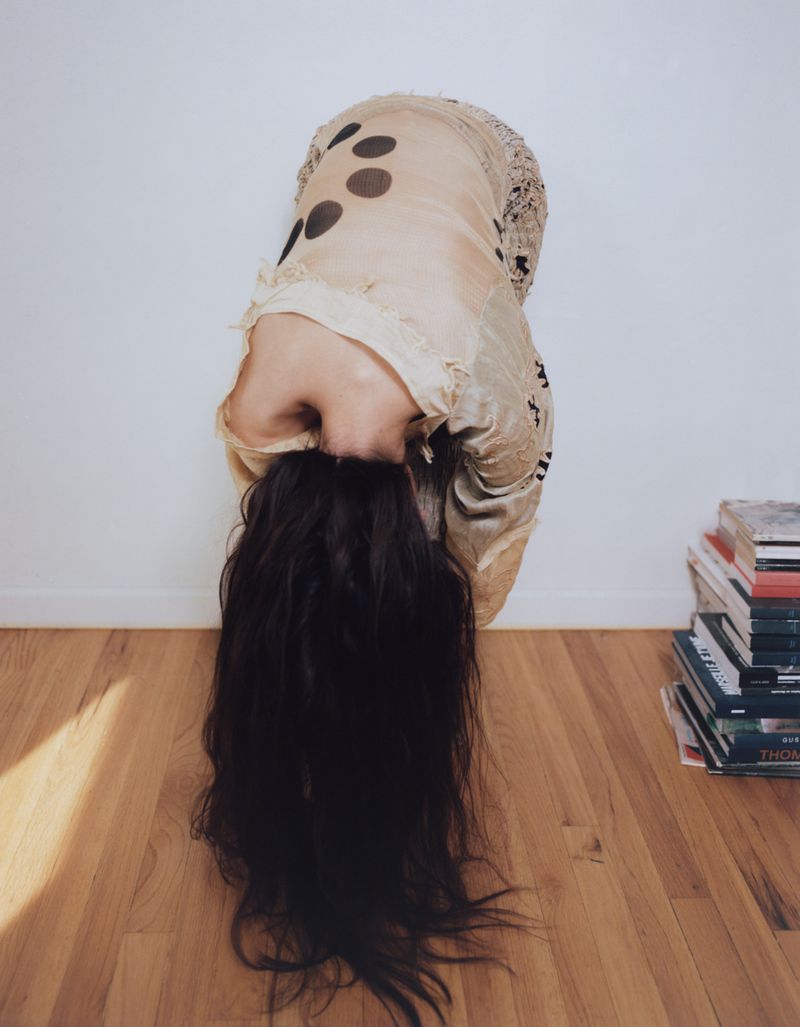 Model bent over forward with dark hair cascading down in front of a white wall and wood floor