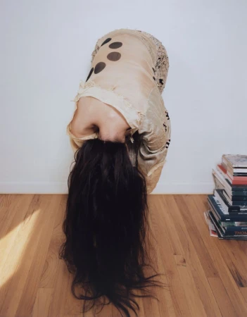 Model bent over forward with dark hair cascading down in front of a white wall and wood floor