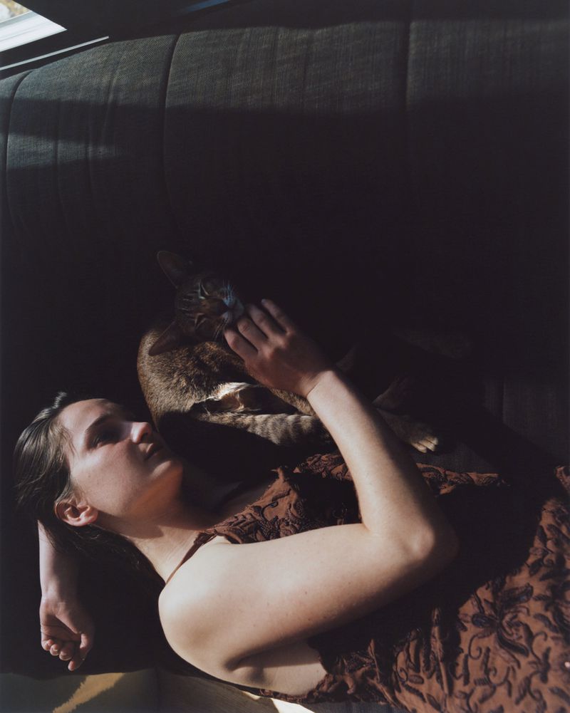 A woman lying on a dark sofa petting a cat resting beside her in warm sunlight.