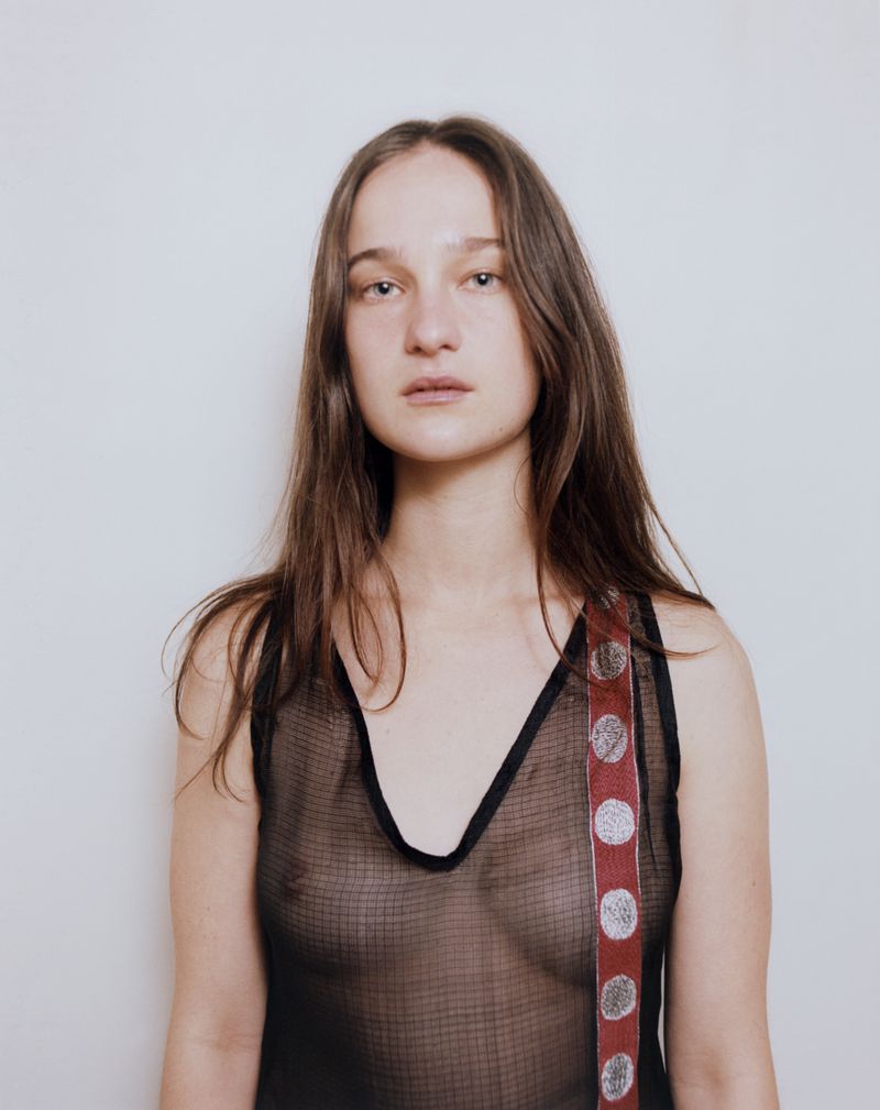 Portrait of a woman wearing a sheer black sleeveless top with a patterned red strap against a plain white wall