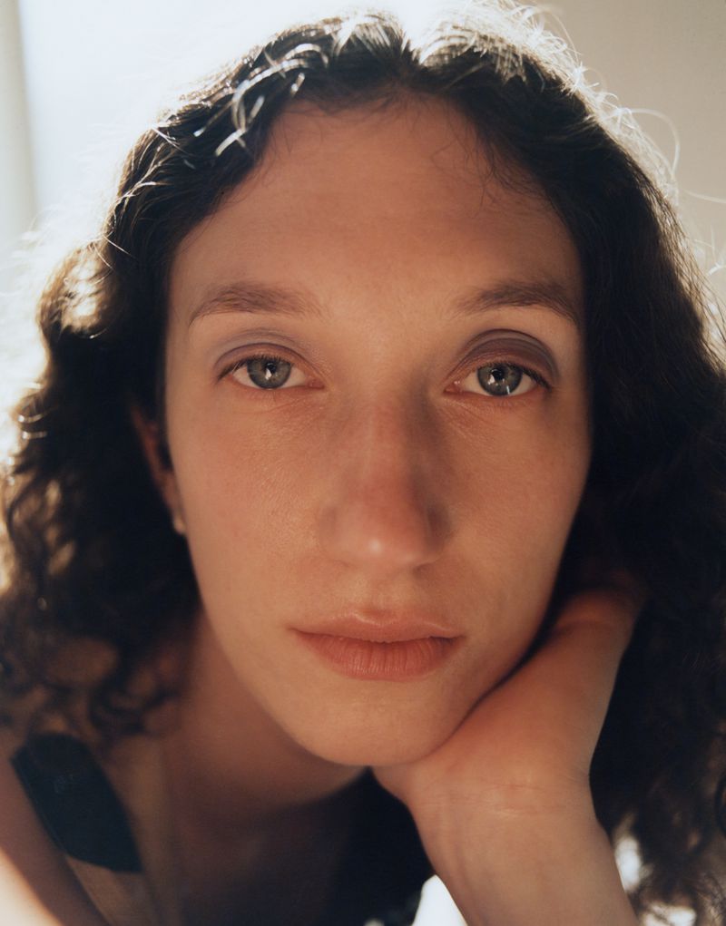 Close-up portrait of a woman with dark curly hair resting her chin on her hand against a bright window light.