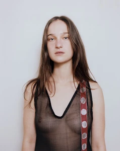 Portrait of a woman wearing a sheer black sleeveless top with a patterned red strap against a plain white wall