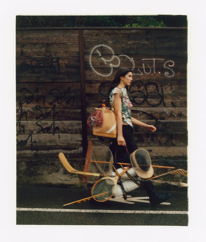Model walking past a graffiti-tagged wooden fence carrying sports equipment and a large tan backpack