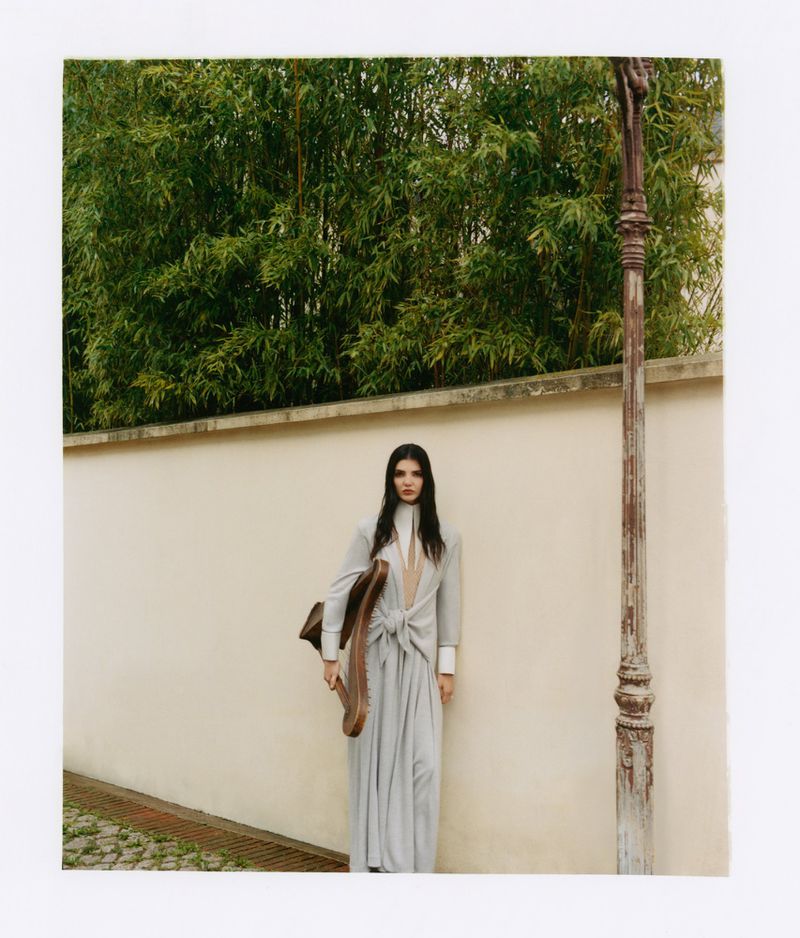 Woman in long grey belted dress holding a harp prop standing against a light wall with bamboo background