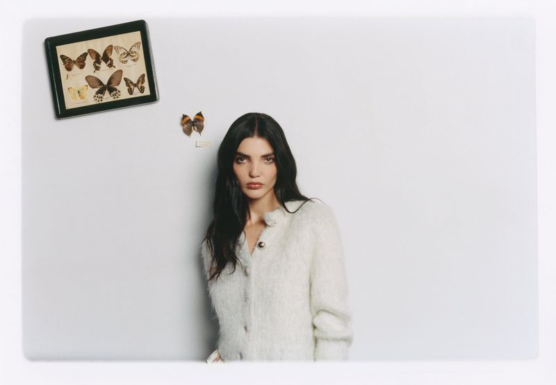 Woman with long dark hair wearing a textured white cardigan posing against a light grey wall with butterfly specimens
