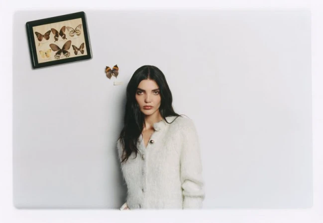 Woman with long dark hair wearing a textured white cardigan posing against a light grey wall with butterfly specimens