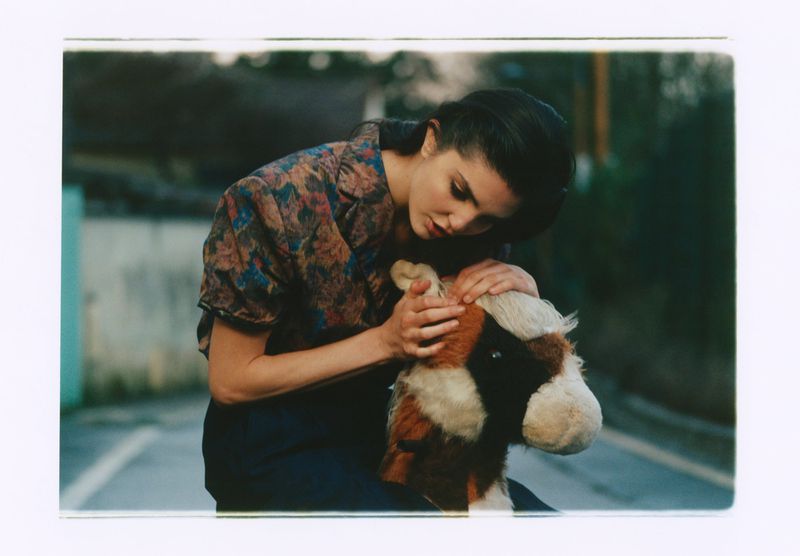 A woman in a floral short-sleeve shirt cradles a stuffed animal horse outdoors against an asphalt background.
