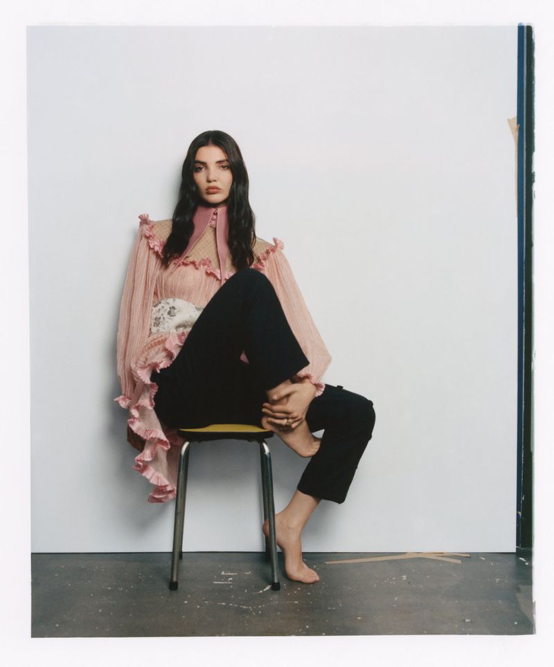 Woman with long dark hair wearing a ruffled pink blouse and black trousers sitting on a small metal stool.