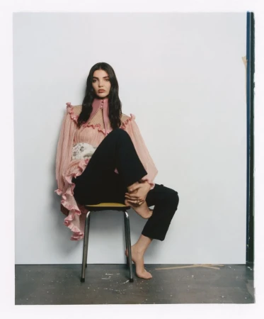 Woman with long dark hair wearing a ruffled pink blouse and black trousers sitting on a small metal stool.