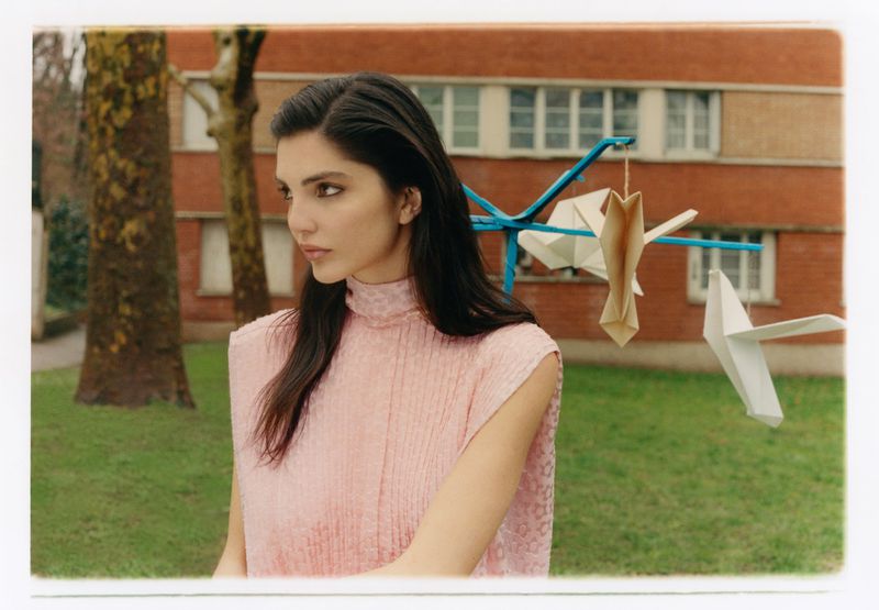 Woman in sleeveless pink textured top outdoors with origami paper birds hanging on a blue metal mobile structure
