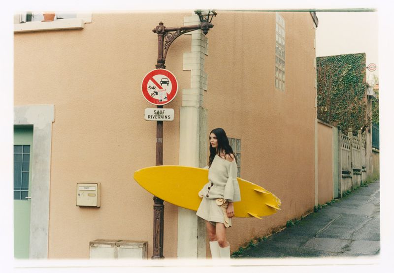 A brunette woman poses on a sidewalk next to a salmon-colored building and street sign while holding a yellow surfboard under her arm.