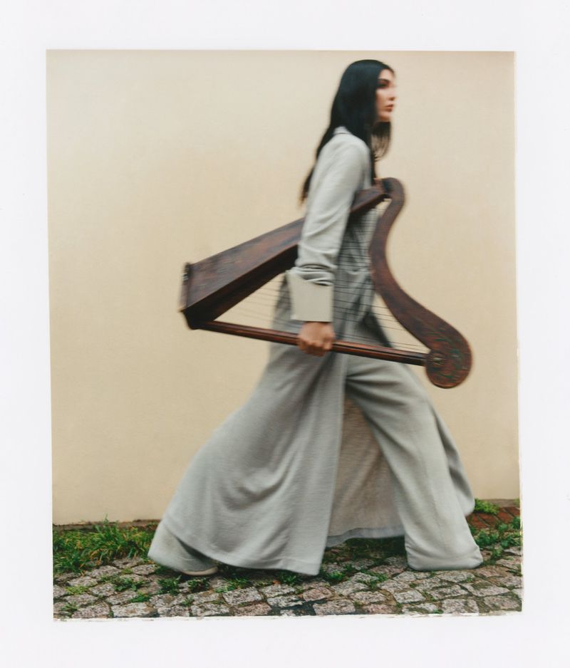 Woman walking outdoors carrying a wooden harp while wearing a long grey coat against a plain wall.