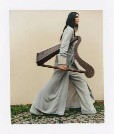 Woman walking outdoors carrying a wooden harp while wearing a long grey coat against a plain wall.