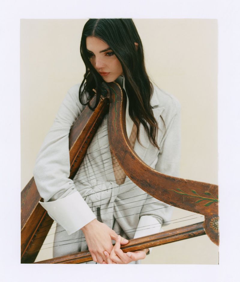 Woman holding a wooden harp wearing a light-coloured blazer against a neutral off-white background.