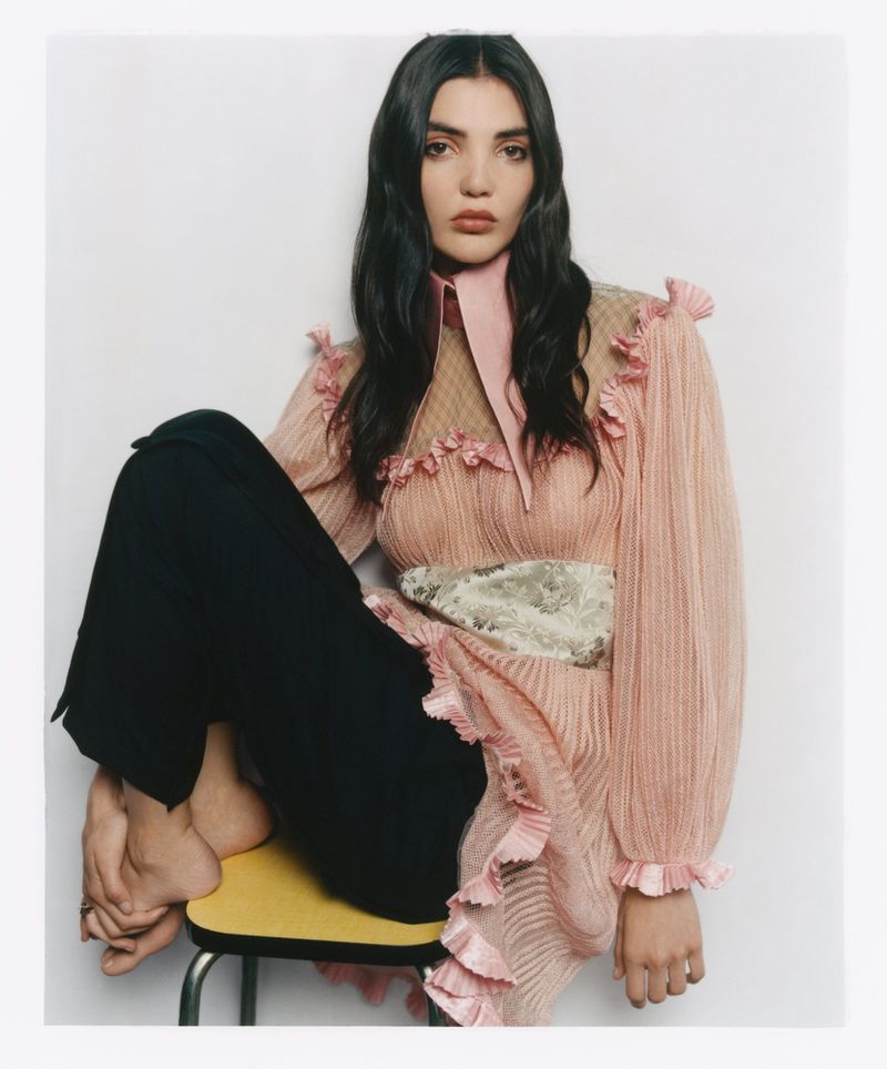 Woman with long dark hair wearing a pink ruffled top and black trousers seated on a yellow stool against a white backdrop