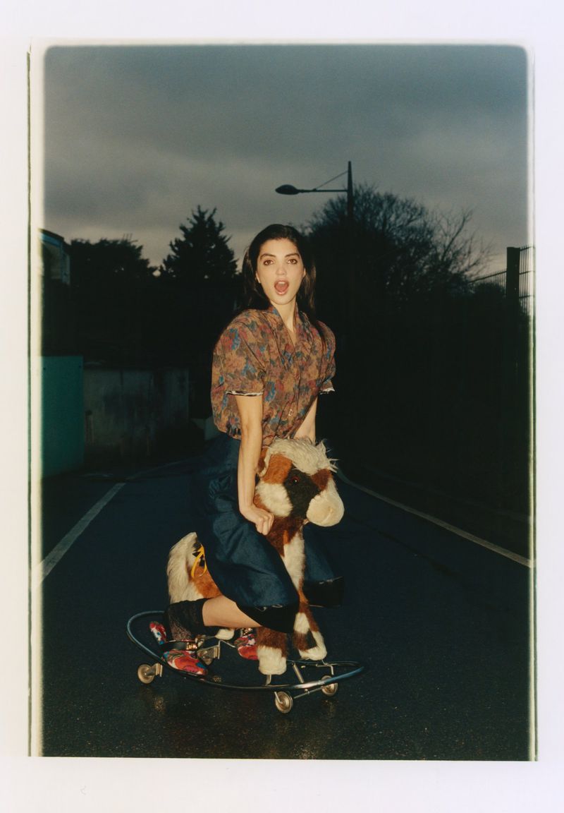 Woman riding a toy rocking horse mounted on a skateboard outdoors on an asphalt road during twilight.