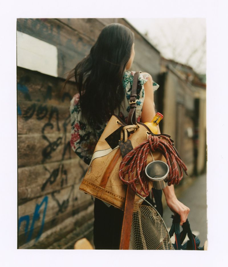 Woman with long dark hair carrying a large patterned handbag and outdoor gear past a weathered wall.