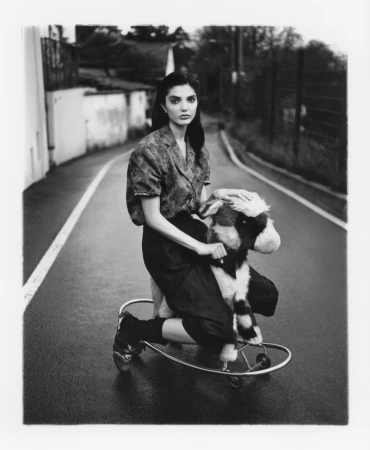 Monochrome fashion editorial image featuring a model riding a wheeled plush animal toy down an empty street.