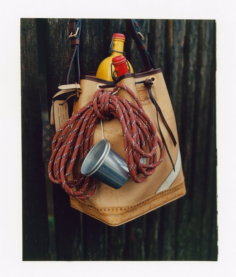 Tan leather bucket bag hanging against a dark wooden slat wall with ropes, a metal cup, and bottles inside.