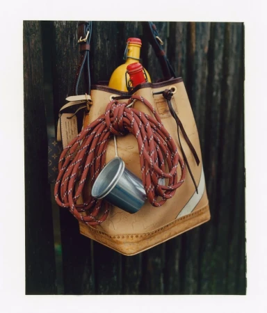 Tan leather bucket bag hanging against a dark wooden slat wall with ropes, a metal cup, and bottles inside.