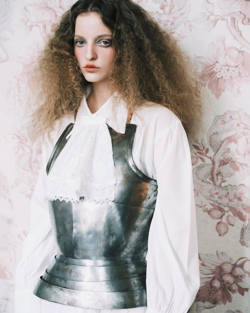 Model wearing a metallic breastplate over a white blouse against a floral patterned wallpaper background