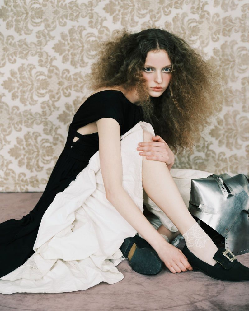 Model with voluminous curly hair seated in a black dress against a damask wallpaper background with a metallic helmet nearby