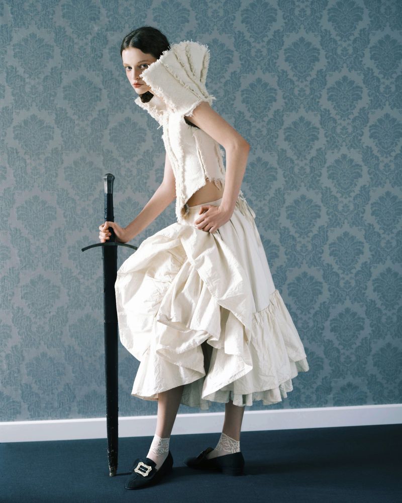 Model in ruffled white top and skirt leaning on a large sword against a patterned blue damask wallpaper for Vogue Portugal.