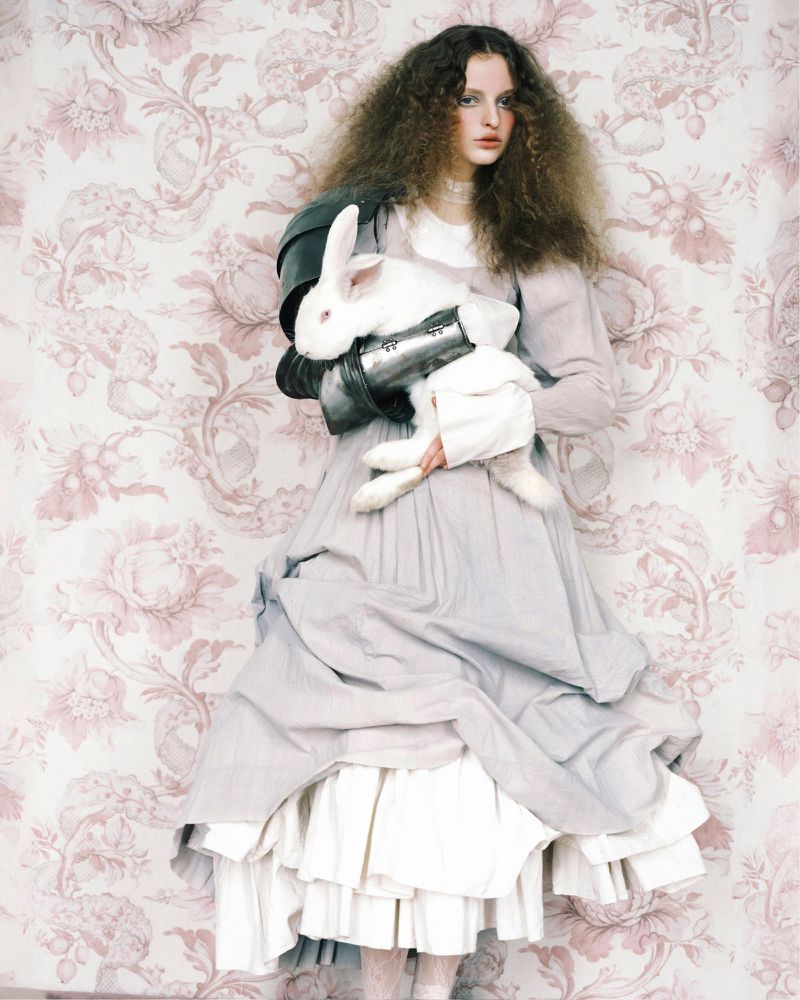 Model with voluminous curly hair wearing a layered grey dress and metal shoulder armor holding a white rabbit