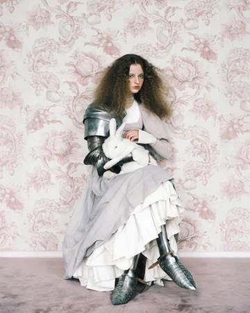 Model wearing medieval metal armour and layered gown holding a white rabbit against pink floral wallpaper for Vogue Portugal.