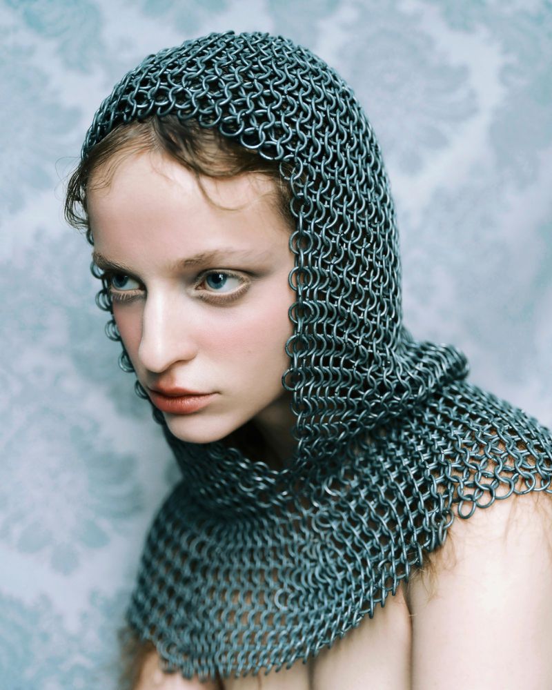 Close-up portrait of a woman wearing a metallic chainmail hood against a patterned blue background.