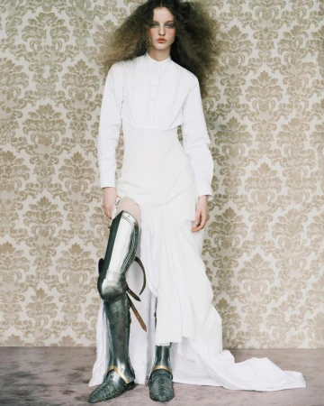 Model in white shirt and skirt wearing metallic greaves against a damask patterned wall