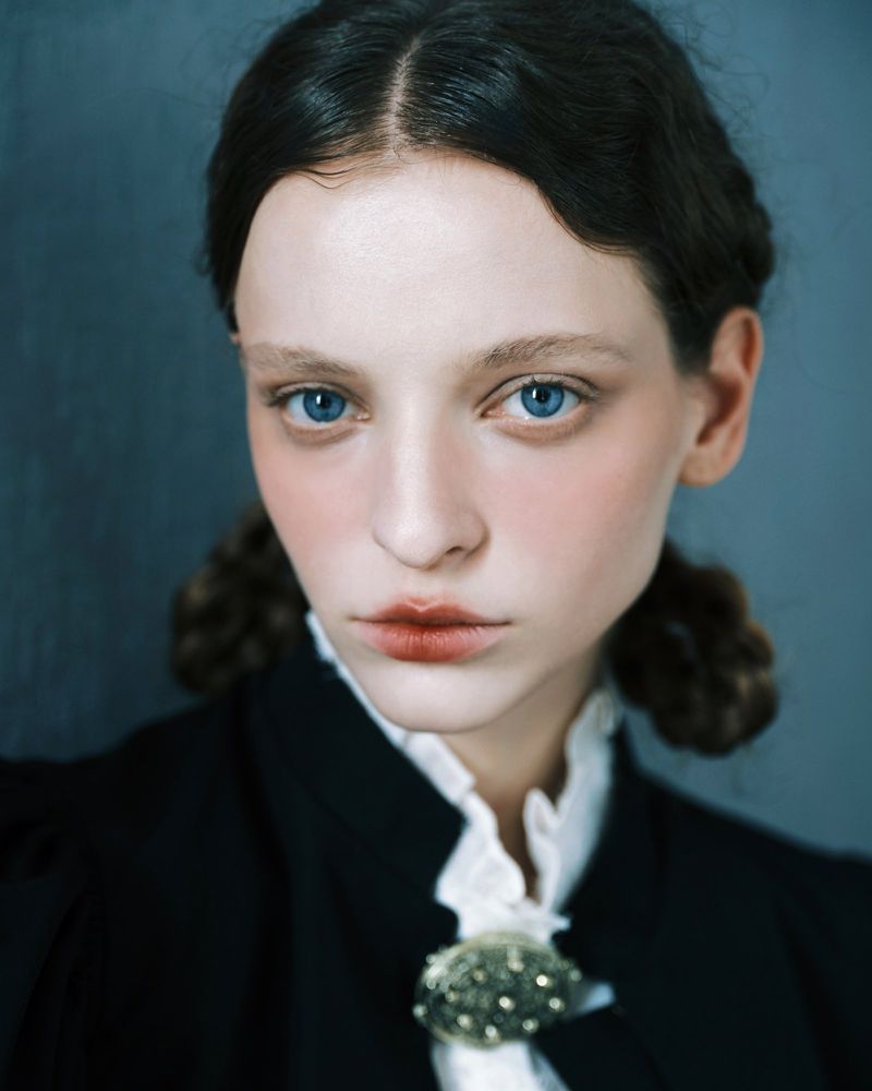 Close up portrait of a young woman with braided dark hair, blue eyes, wearing a dark jacket with a white ruffled collar.