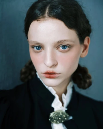 Close up portrait of a young woman with braided dark hair, blue eyes, wearing a dark jacket with a white ruffled collar.