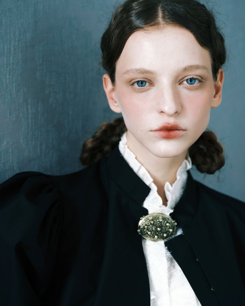 Portrait of a female model in a black jacket and white ruffled blouse with an ornate brooch against a textured grey wall