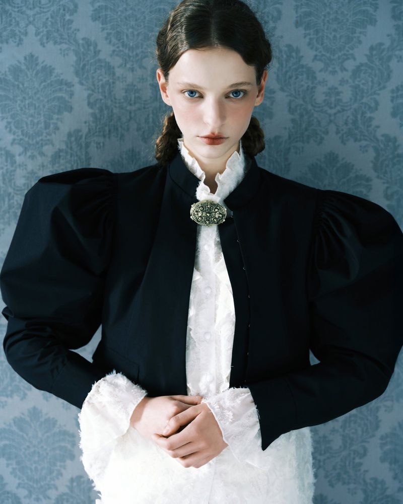 Model wearing a structured black jacket over a white ruffled blouse against a blue damask wallpaper background
