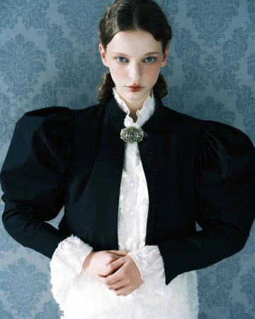 Model wearing a structured black jacket over a white ruffled blouse against a blue damask wallpaper background
