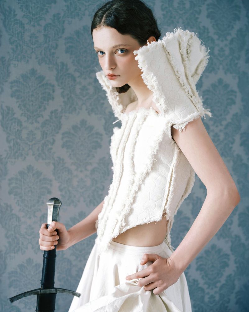 Model wearing a white structured top with frayed edges holding a sword against a patterned blue wallpaper background