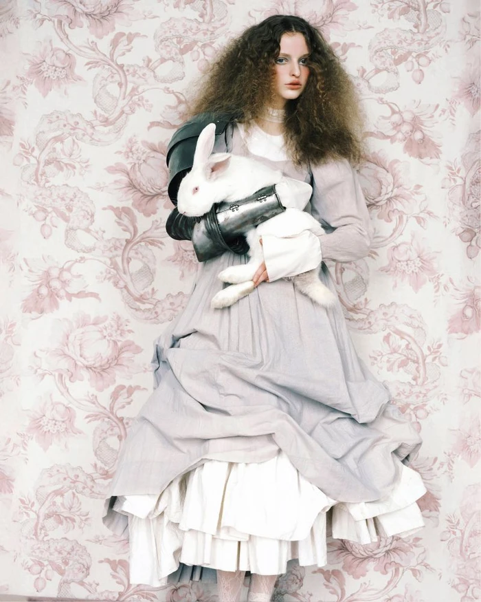 Model with voluminous curly hair wearing a layered grey dress and metal shoulder armor holding a white rabbit