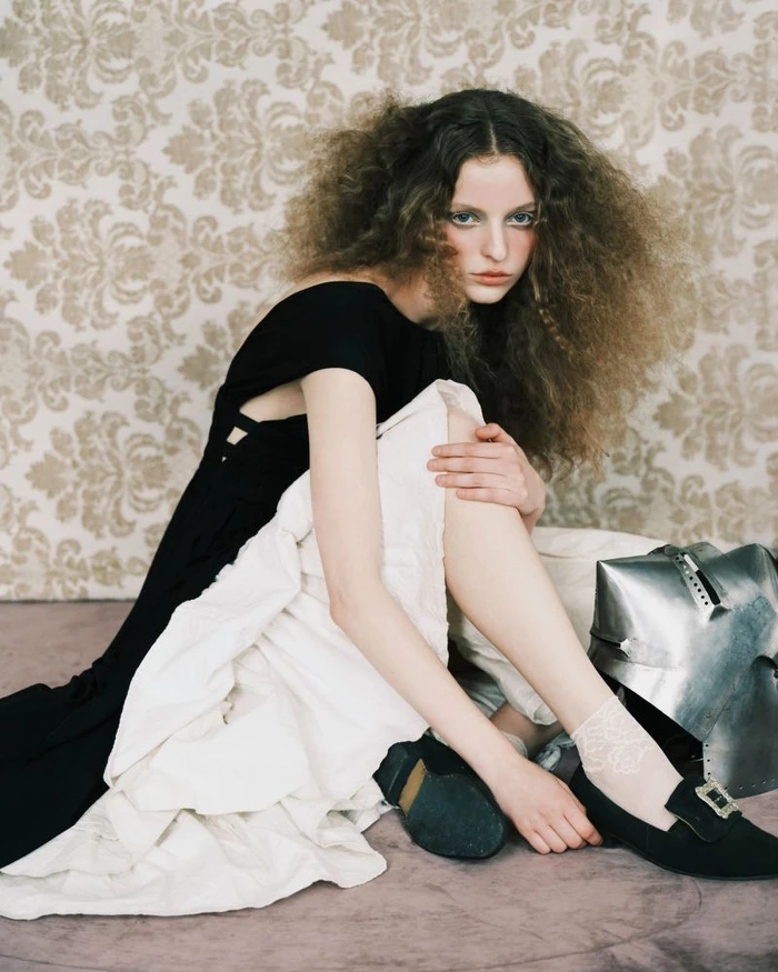 Model with voluminous curly hair seated in a black dress against a damask wallpaper background with a metallic helmet nearby