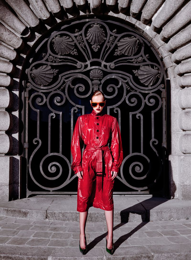 Model in shiny red coat and sunglasses standing before an ornate wrought-iron gate for V Magazine China editorial by Yis Kid
