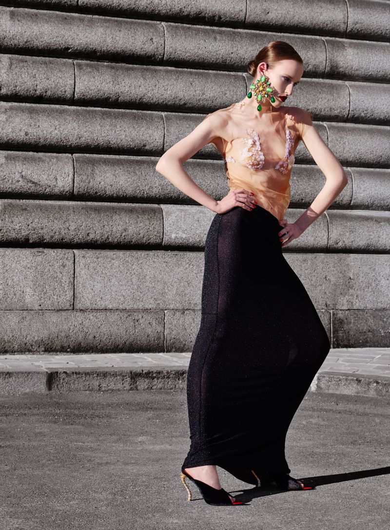Fashion shot of a woman in a sheer top and long skirt posing in front of gray stone steps wearing statement earrings and black mules.