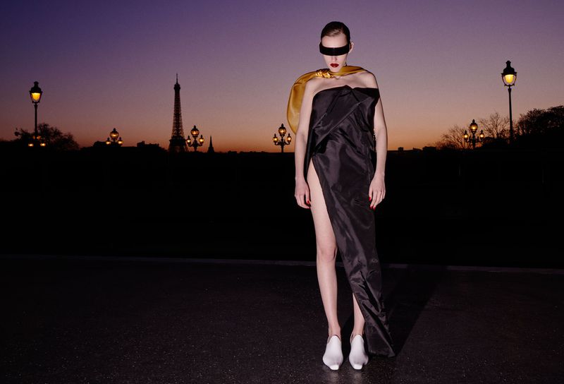 Model standing outdoors at dusk wearing a strapless black dress, gold cape, white shoes, and a black visor over eyes.
