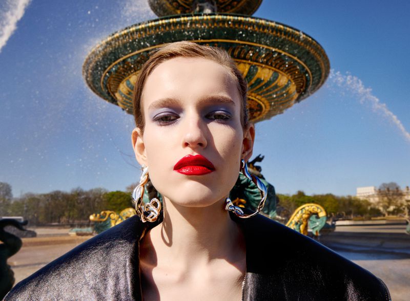 Close-up portrait of a woman wearing glossy red lipstick, blue eye shadow, and large silver earrings in front of a classical fountain.