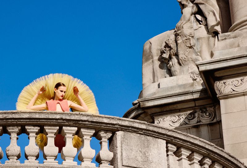 Model wearing a pleated pink top with a large yellow tulle ruff standing on a stone balcony against a clear blue sky